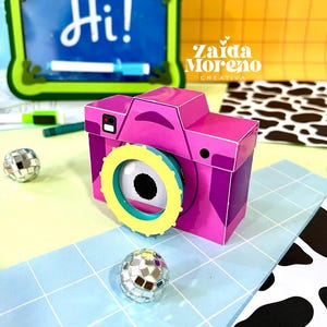 Puede incluir: Una cámara de papel tridimensional en tonos magenta y morado, con un anillo de lente amarillo brillante y un gráfico de ojo en blanco y negro en el centro. La cámara está colocada sobre una superficie de cuadrícula azul claro con dos bolas de discoteca.