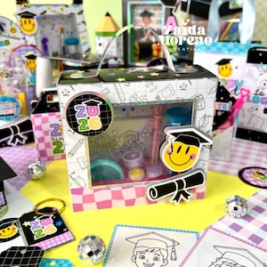 May include: A square cardboard gift box with a clear window, featuring a school-themed pattern, a handle, and a circular tag with the text 2026. The box is decorated with a smiley face wearing a graduation cap and a diploma icon, surrounded by coloring sheets.