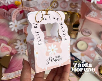 May include: A pink and white gift box with a handle and a daisy pattern. The box has the text "Feliz Día de las Madres" and "Te Amo Mamá" on it. The box is sitting on a hand.