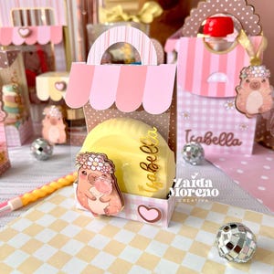 May include: A small pink and white striped paper favor box with a scalloped top handle. The box features a cartoon animal wearing a flower crown, a yellow circular item, and the name Isabella in gold script. The box sits on a checkered surface.
