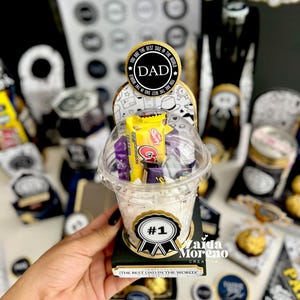 May include: A clear plastic cup filled with candy, topped with a lid, and set on a black base. The cup has a sticker that says "#1" and "The Best Dad in the World!". A circular sticker on top says "DAD".
