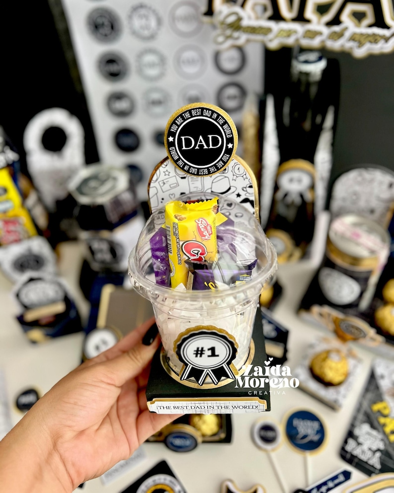 May include: A clear plastic cup filled with candy, topped with a lid, and set on a black base. The cup has a sticker that says "#1" and "The Best Dad in the World!". A circular sticker on top says "DAD".