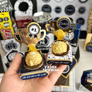 May include: Gold-colored trophy-shaped gift boxes with a chocolate candy inside. One trophy has a "#1" ribbon, the other has a "DAD" medallion. The boxes have text that reads "The best dad in the world!" and "El mejor del mundo!"
