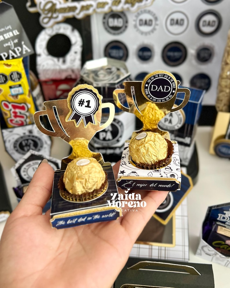 May include: Gold-colored trophy-shaped gift boxes with a chocolate candy inside. One trophy has a "#1" ribbon, the other has a "DAD" medallion. The boxes have text that reads "The best dad in the world!" and "El mejor del mundo!"