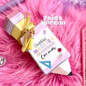 Può includere: Una confezione regalo a forma di matita con un nastro dorato, una stella e un nappa. La scatola è gialla con una parte superiore glitterata rosa e una carta a forma di matita con la scritta "Gracias Maestra por enseñarme con amor". Lo sfondo è rosa.