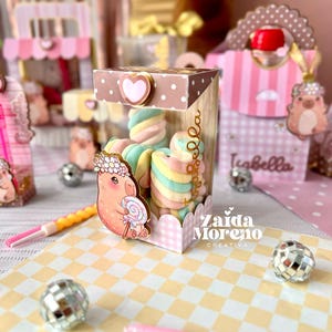 Puede incluir: Una caja de cartón rectangular con ventana transparente llena de malvaviscos trenzados de colores pastel. La caja presenta una tapa marrón con lunares blancos, un emblema de corazón y un personaje de dibujos animados con una piruleta. Texto Zaida Moreno.