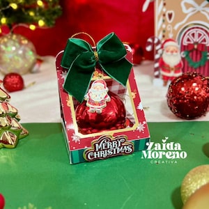 Puede incluir: Un adorno navideño en una caja decorativa con un lazo verde. La caja tiene las palabras "Merry Christmas" y un adorno de Papá Noel. Otras decoraciones navideñas incluyen un árbol dorado y adornos rojos.