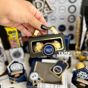 May include: A small, black gift box with a gold handle, containing two gold-wrapped chocolate candies. A round sticker with the text "PARA EL MEJOR PAP&Aacute;" is on the box. Other gift boxes are in the background.