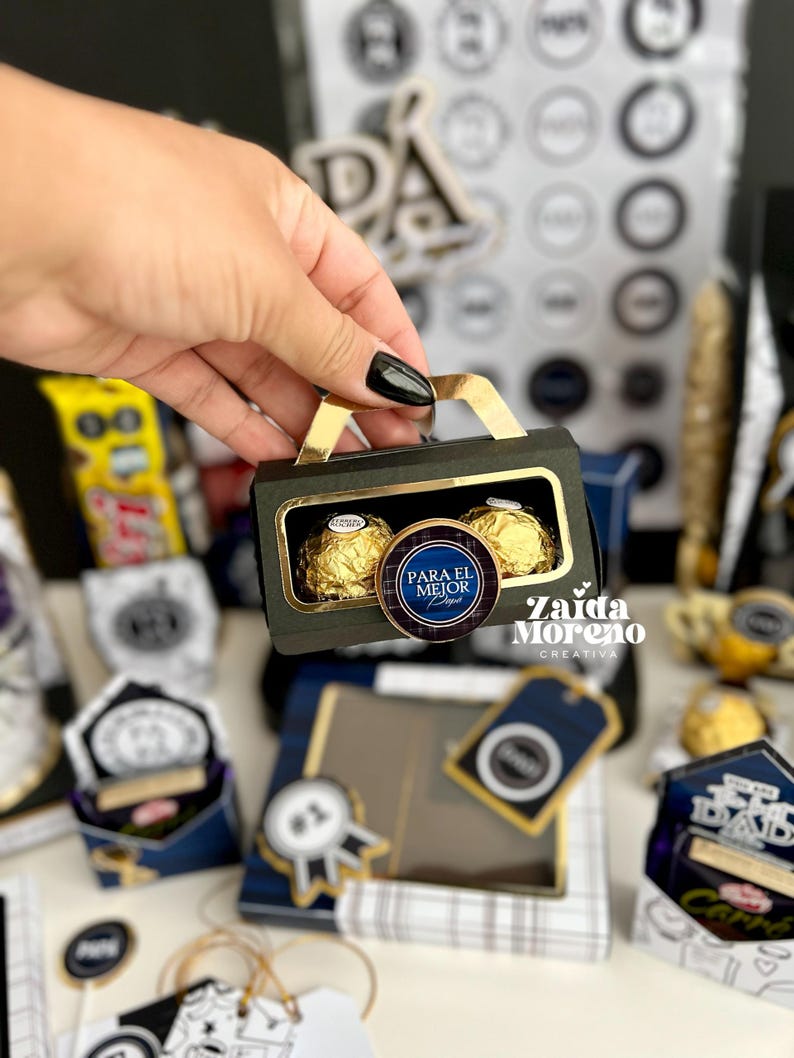 May include: A small, black gift box with a gold handle, containing two gold-wrapped chocolate candies. A round sticker with the text "PARA EL MEJOR PAP&Aacute;" is on the box. Other gift boxes are in the background.