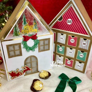 Puede incluir: Dos calendarios de adviento con forma de casa y borde dorado. Uno tiene una escena navideña con una corona y el texto "Merry Christmas". El otro tiene cajones numerados. Chocolates dorados y un lazo verde en primer plano.