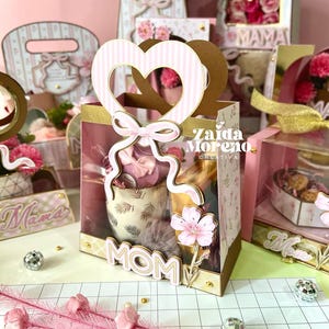 May include: A collection of decorative gift boxes and bags in shades of pink, white, and gold. The central box features a heart-shaped handle and the word "MOM". Other boxes include floral and striped patterns, bows, and the word "MAMA".