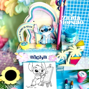 Puede incluir: Un colorido kit de manualidades con el personaje Stitch de Lilo & Stitch. El kit incluye una figura de Stitch, botes de pintura, lápices y una página para colorear. Se ven las palabras "aloha" y "Llena tu mundo de color".