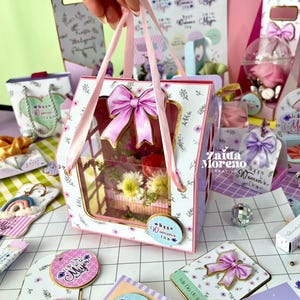Pode incluir: Uma caixa de presente floral branca e rosa com um grande laço rosa e uma janela transparente, cheia de flores. A caixa tem alças de fita rosa e é cercada por outros itens de presente com desenhos florais e o texto "Happy Women's Day".