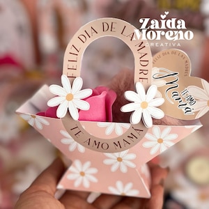 Puede incluir: Una cesta de papel rosa y blanca con un patrón de margaritas. La cesta tiene un asa con el texto "Feliz Dia de la Madre" y una etiqueta en forma de corazón con el texto "Te Amo Mamá".