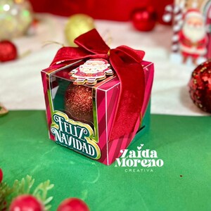 Könnte beinhalten: Eine festliche Weihnachtsgeschenkbox mit einer roten Samtschleife und einem Weihnachtsmann-Ornament. Die Box ist rosa und grün mit den Worten "Feliz Navidad" auf der Vorderseite. Im Inneren befindet sich ein roter Glitzer-Ornament. Weihnachtsdekoration.