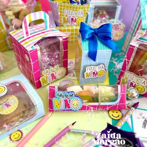 Peut inclure: Une collection de bo&icirc;tes cadeaux et de contenants color&eacute;s, certains avec des fen&ecirc;tres transparentes, remplis de friandises. Les bo&icirc;tes sont d&eacute;cor&eacute;es de rayures, de n&oelig;uds et du texte "Feliz D&iacute;a del Ni&ntilde;o" et un soleil souriant. Des crayons et des autocollants smiley sont &eacute;galement visibles.