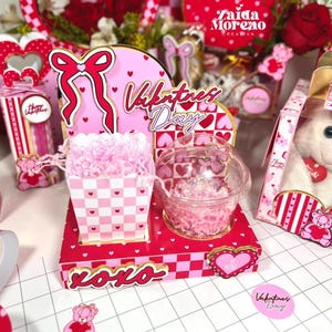 Puede incluir: Una exhibición del Día de San Valentín con un esquema de color rojo y rosa. La exhibición incluye una caja con un patrón de cuadros rosas y blancos, un recipiente transparente y elementos decorativos con corazones y los textos "XOXO" y "Valentines Day".