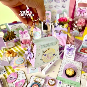 Puede incluir: Una colección de cajas de regalo, etiquetas y tarjetas con detalles florales y lazos. La caja principal es azul claro con un asa dorada y el texto "Feliz Dia de la Mujer". Otros artículos incluyen un llavero arcoíris y una tarjeta con un chocolate.