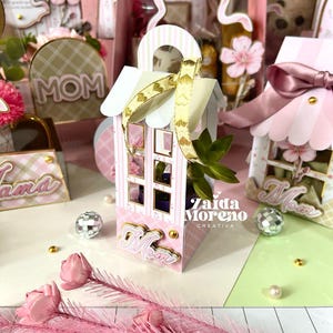 Könnte beinhalten: Rosa und weiße dekorative Geschenkboxen mit floralen Akzenten und dem Wort „Mom“. Die Boxen haben ein hausähnliches Design mit Griff und goldenem Band. Das Bild enthält auch dekorative Blumen und Discokugeln.