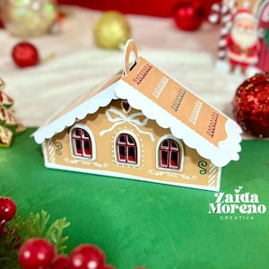 May include: A gingerbread house-shaped ornament with a tan exterior, white frosting details, and red window frames. The roof has white and tan accents. The ornament is on a green surface with Christmas decorations in the background.