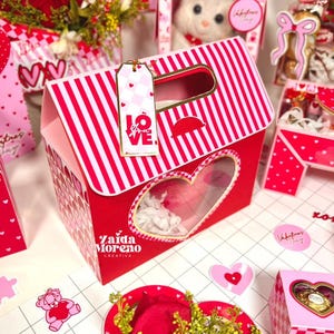 Puede incluir: Caja de regalo a rayas rojas y blancas con ventana en forma de corazón, asa dorada y etiqueta con la palabra "LOVE". La caja forma parte de una temática de San Valentín, con otros regalos y decoraciones con forma de corazón.