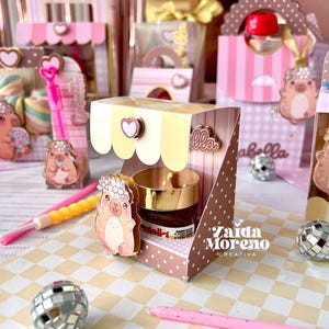 Puede incluir: Un pequeño tarro de Nutella en una caja de presentación de papel rosa y crema con un toldo festoneado, un corazón decorativo y un personaje de dibujos animados con corona de flores. La caja incluye el texto Zaida Moreno CREATIVA y lunares.