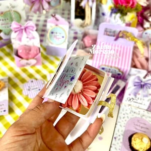 Pode incluir: Uma mão segura uma pequena caixa de presente em forma de livro aberta com uma margarida rosa dentro. A caixa tem detalhes dourados e o texto "Feliz Dia de la Mujer". Outros presentes com laços e etiquetas estão no fundo.