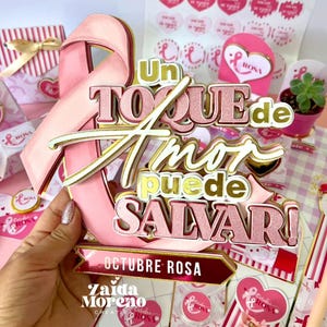 以下が含まれることがあります： ピンクのリボンのケーキトッパー、ゴールドのアクセントと「Un Toque de Amor puede SALVAR！」の文字がピンクとゴールドで書かれています。「OCTUBRE ROSA」の文字も表示されています。他のピンクと白の装飾品が背景にあります。