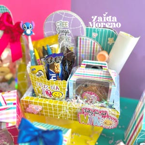 Peut inclure: Un panier cadeau color&eacute; rempli de friandises et de fournitures scolaires. Le panier comprend une bo&icirc;te de biscuits, des bonbons, des crayons et un stylo avec un personnage Stitch. Les mots "Feliz D&iacute;a del Ni&ntilde;o" sont visibles.