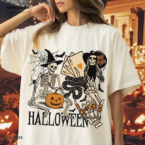 May include: Off-white Halloween-themed t-shirt with a graphic featuring skeletons, playing cards, a jack-o'-lantern, and the word "HALLOWEEN". The design includes bats, a spider web, and the text "HOWDY HALLOWEEN".