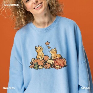 May include: A light blue crewneck sweatshirt featuring a graphic of two orange kittens sitting on pumpkins surrounded by autumn leaves. A red circular stamp with the text MONOLINES is overlaid on the right side of the graphic.