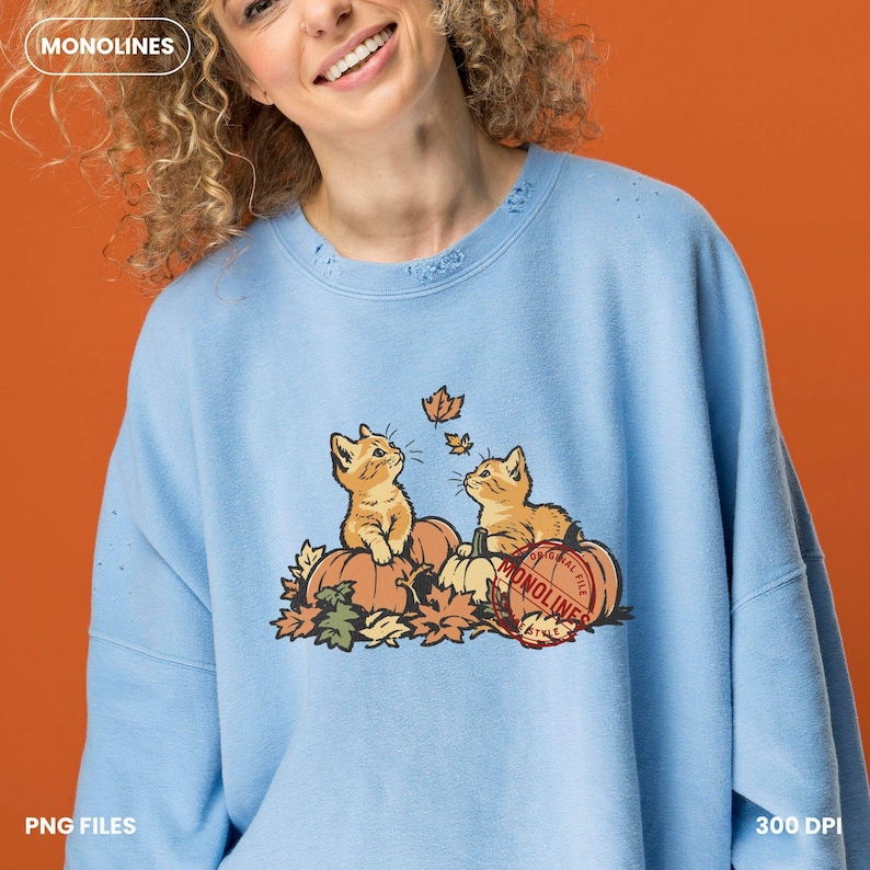 May include: A light blue crewneck sweatshirt featuring a graphic of two orange kittens sitting on pumpkins surrounded by autumn leaves. A red circular stamp with the text MONOLINES is overlaid on the right side of the graphic.