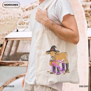 May include: A beige canvas tote bag featuring a graphic illustration of a tan calf wearing a black witch hat and black boots, holding a small orange pumpkin bucket. The design includes a purple circular stamp overlay and scattered leaves.