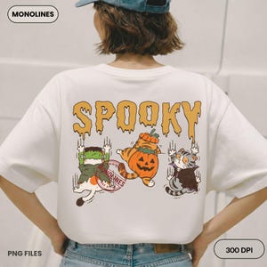 May include: A white t-shirt featuring a graphic on the back with the text SPOOKY in orange dripping letters. The design includes three cats dressed as a Frankenstein monster, a pumpkin, and a vampire, with claw marks and a red circular stamp.