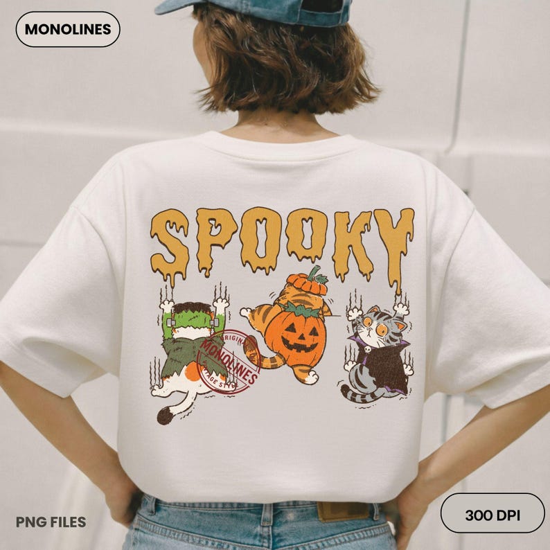 May include: A white t-shirt featuring a graphic on the back with the text SPOOKY in orange dripping letters. The design includes three cats dressed as a Frankenstein monster, a pumpkin, and a vampire, with claw marks and a red circular stamp.