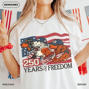 May include: White t-shirt with a patriotic design. It features three dogs wearing bandanas and caps, set against an American flag. The text "250 YEARS OF FREEDOM" is printed below the dogs.