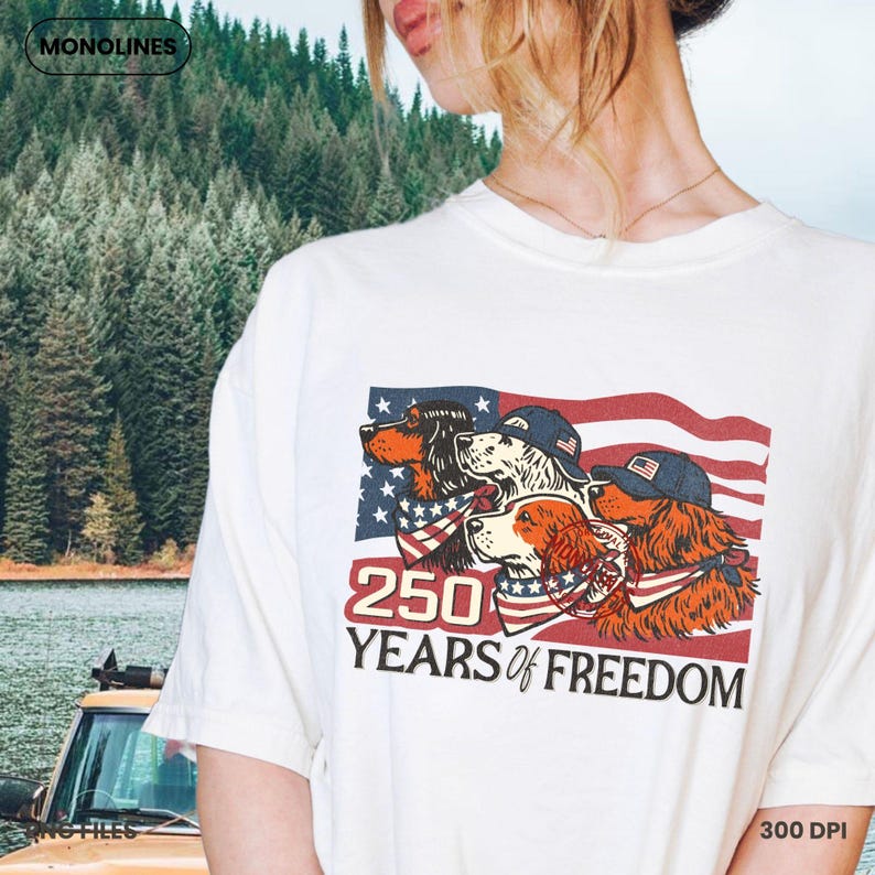 May include: Cream-coloured t-shirt with a graphic of four dogs wearing patriotic bandanas and caps, set against an American flag design. The text "250 YEARS of FREEDOM" is below the graphic. The shirt has a crew neck.