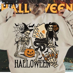 May include: Beige Halloween-themed sweatshirt featuring a skeleton wearing a witch hat, a jack-o'-lantern, playing cards, and the text "Howdy Halloween." The design includes bats and a spider web. The word "Halloween" is printed at the bottom.