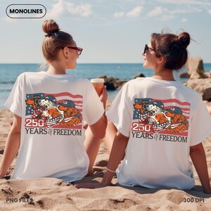 May include: White t-shirts with a graphic of three dogs in patriotic attire, set against an American flag design. The text "250 YEARS OF FREEDOM" is below the graphic. The image is on a beach with two people.