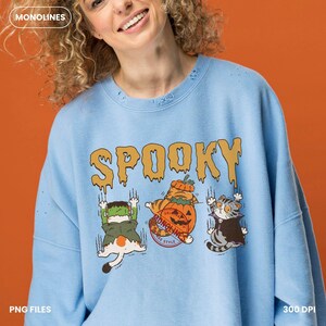 May include: A light blue crewneck sweatshirt featuring the text SPOOKY in a dripping yellow font. Below the text are three illustrated cats: one dressed as Frankenstein, one inside a pumpkin, and one wearing a vampire cape, all with claw marks.