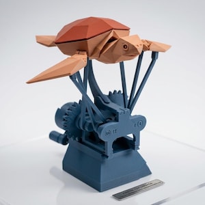 Può includere: Una scultura geometrica stampata in 3D di una tartaruga con guscio marrone e appendici beige. La tartaruga è montata su una base meccanica blu con ingranaggi e strutture di supporto. Una piccola placca metallica è alla base.