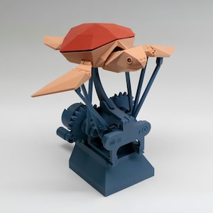 Puede incluir: Una escultura cinética mecánica que presenta una tortuga marina de bajo poligonaje con caparazón rojo y cuerpo color melocotón claro. La tortuga está montada sobre una base azul oscuro con un complejo sistema de engranajes, palancas y brazos.