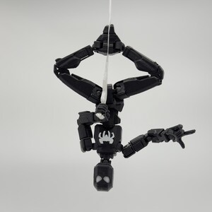 May include: A black articulated action figure of a superhero, hanging upside down from a white string. The figure has a spider emblem on its chest and a mask with white eye details. The figure is posed as if shooting a web.