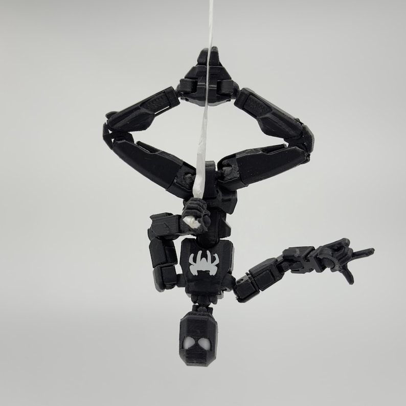 May include: A black articulated action figure of a superhero, hanging upside down from a white string. The figure has a spider emblem on its chest and a mask with white eye details. The figure is posed as if shooting a web.
