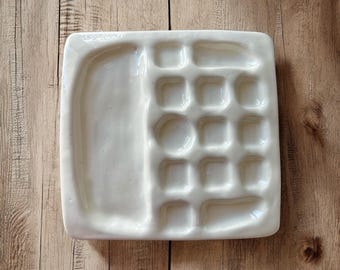 May include: White ceramic paint palette with a large well and a grid of smaller wells.