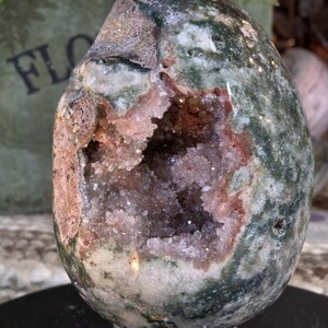 May include: An egg-shaped stone geode featuring a polished exterior with mottled dark green, white, and brown patterns. The center of the stone is open, revealing a hollow cavity lined with small, sparkling, light pink and translucent crystals.