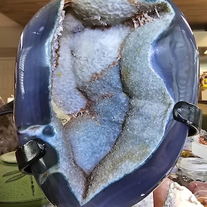 May include: A polished blue agate geode with a large, irregular central cavity filled with sparkling, light-colored crystalline formations. The outer surface of the stone is smooth and deep blue, held in place by a black metal display stand.