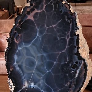 May include: A large, polished agate slice featuring deep navy blue and black tones with intricate, translucent white veining that creates a web-like pattern. The outer edge of the stone is rough, textured, and colored in shades of tan and cream.