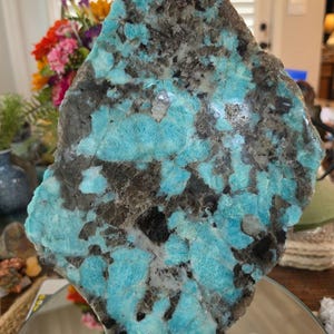 May include: A large, flat, irregularly shaped slab of polished mineral featuring a vibrant mosaic of bright turquoise blue and dark charcoal gray to black crystalline inclusions. The surface displays a glossy, reflective finish with distinct color patterns.