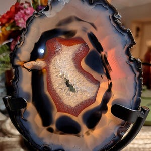 May include: A translucent agate slice featuring concentric bands of dark charcoal and translucent amber. The center contains a crystalline geode cavity with a vibrant orange-red border, all supported by a black metal display stand.